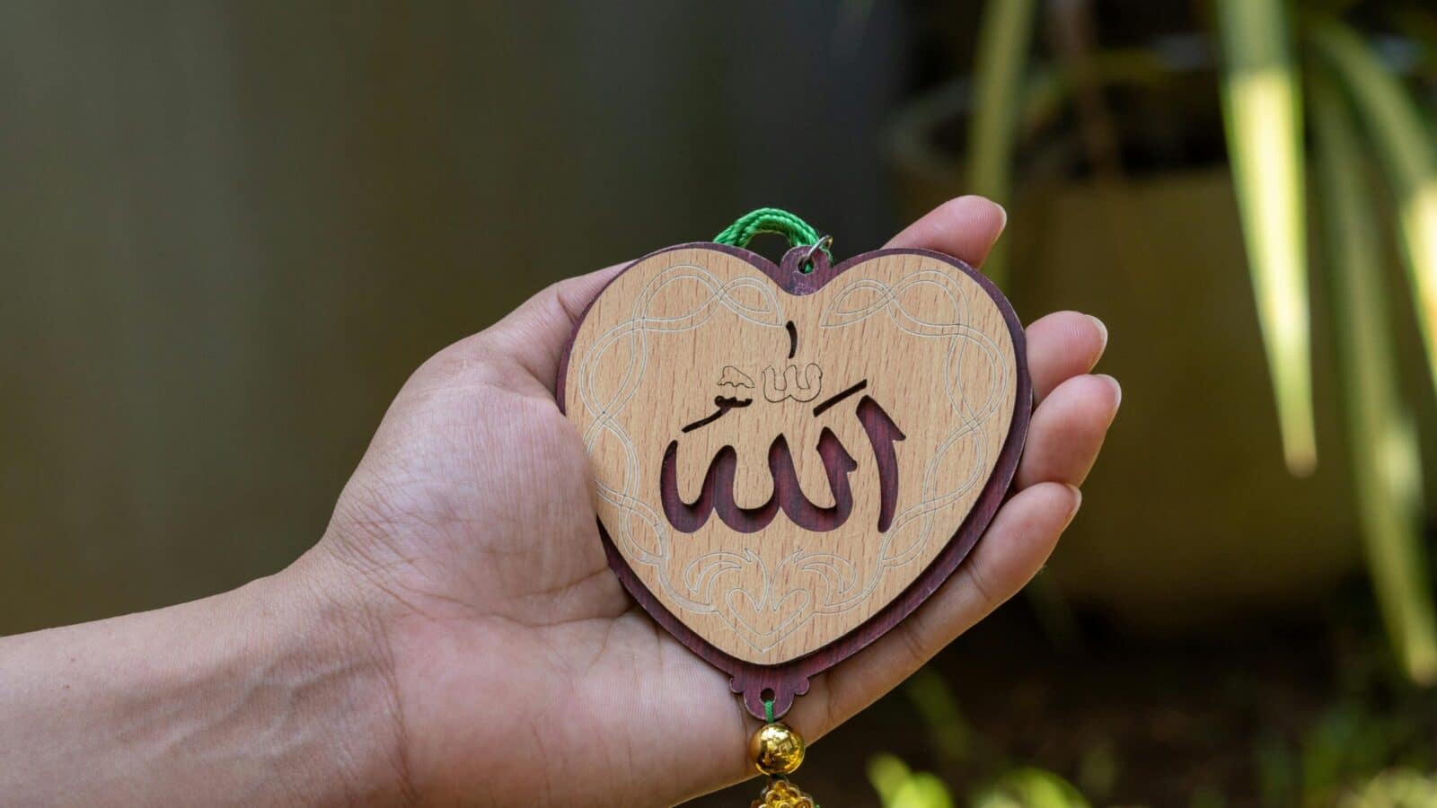 Person holding a heart-shaped wooden piece with Islamic calligraphy outdoors.