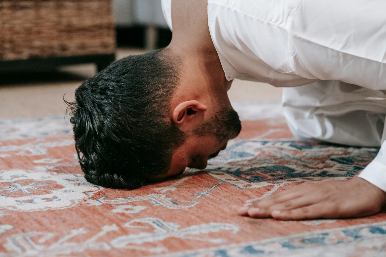A man in a white thobe performs salah, kneeling on a patterned prayer rug indoors.