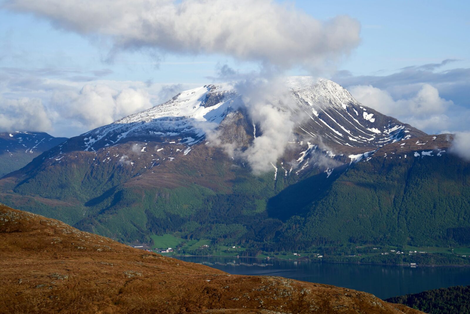 Serene landscape of a snow-capped mountain partially obscured by clouds, overlooking a tranquil lake.