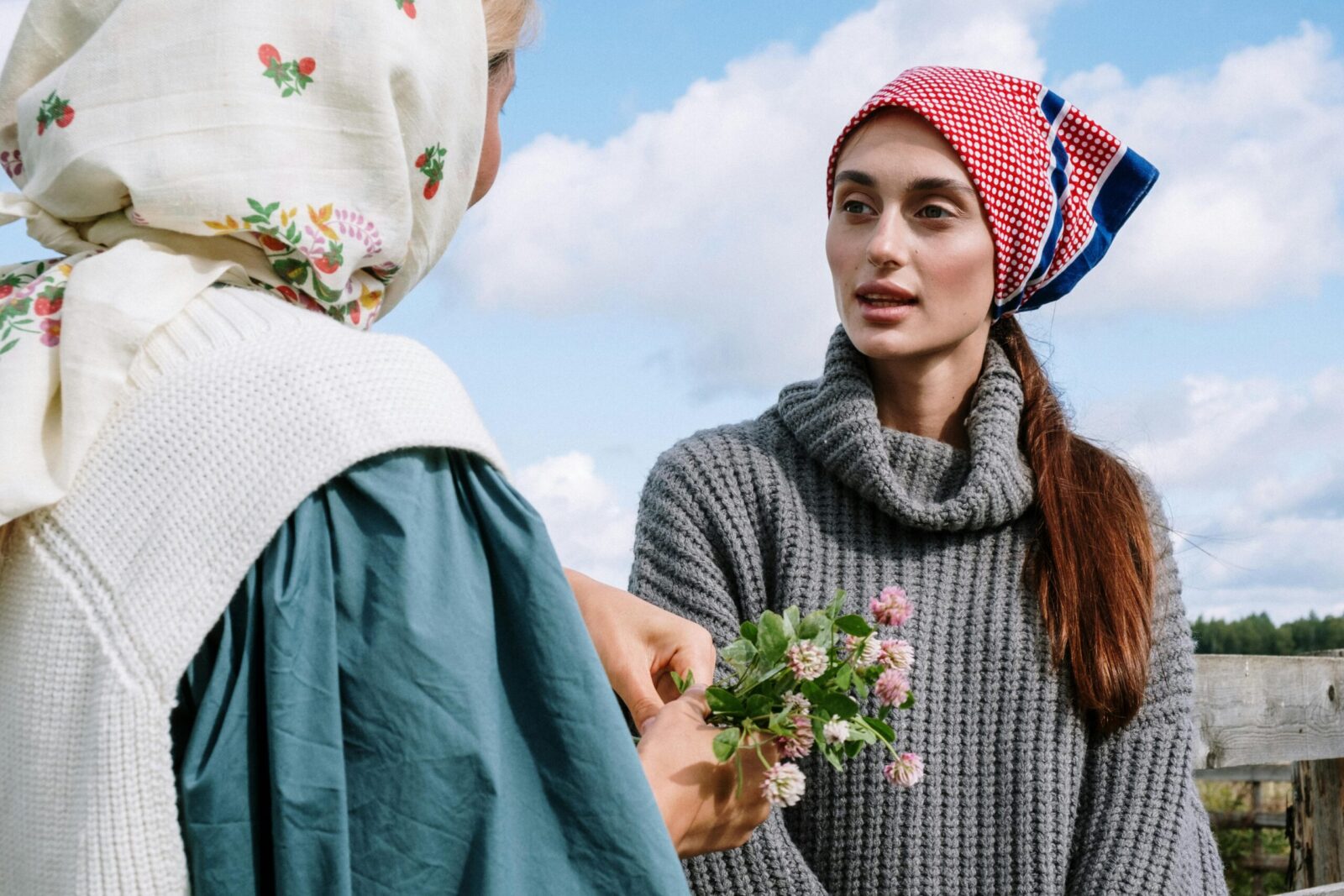 Two women outdoors in headscarves holding wildflowers under a clear sky.