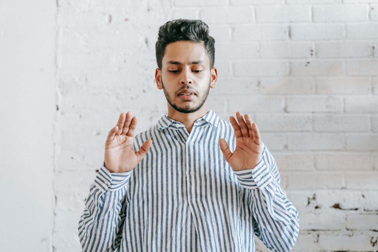 An adult man wearing a striped shirt, engaged in a prayer ritual indoors against a white brick wall.