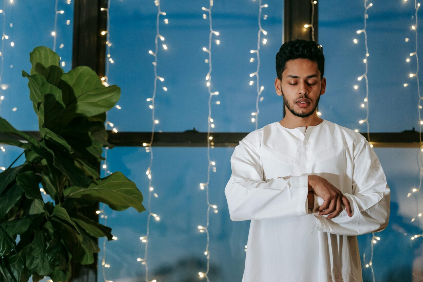 A man wearing traditional attire stands indoors near a window, illuminated by string lights, engaged in prayer.