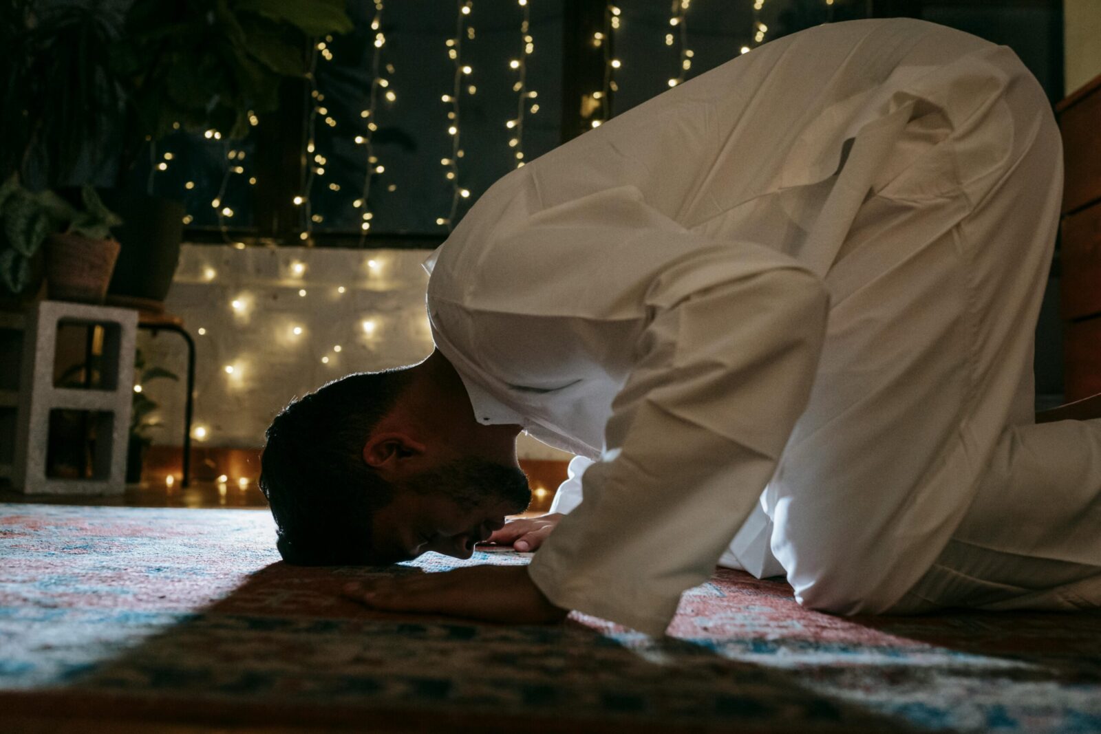 A man in traditional attire bows in prayer on a rug lit by string lights, symbolizing faith and devotion.