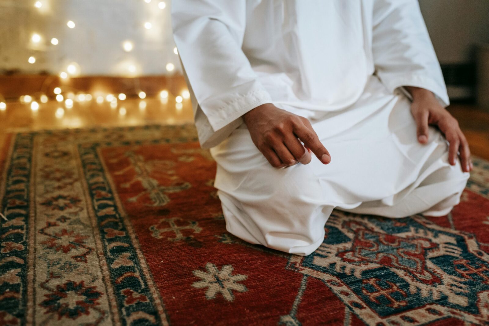 A man in traditional attire kneeling in prayer indoors with string lights overhead.