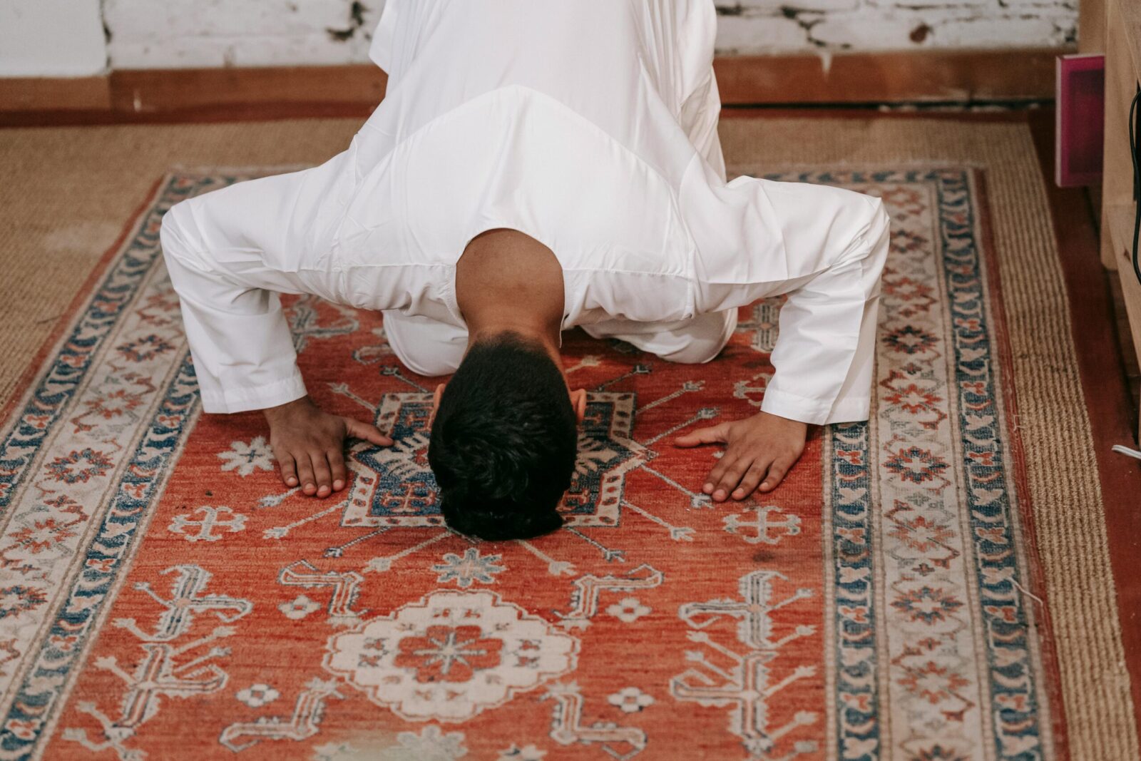 A Muslim man in traditional clothing performs salah on a beautifully designed prayer rug inside a room.