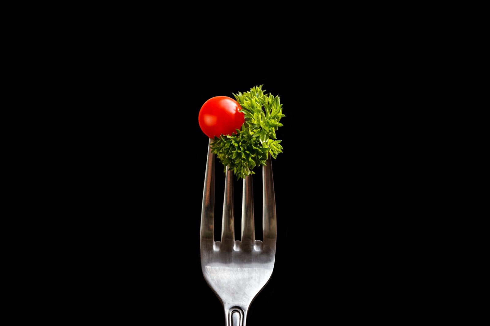 Close-up of a fork with cherry tomato and parsley against a dark background.