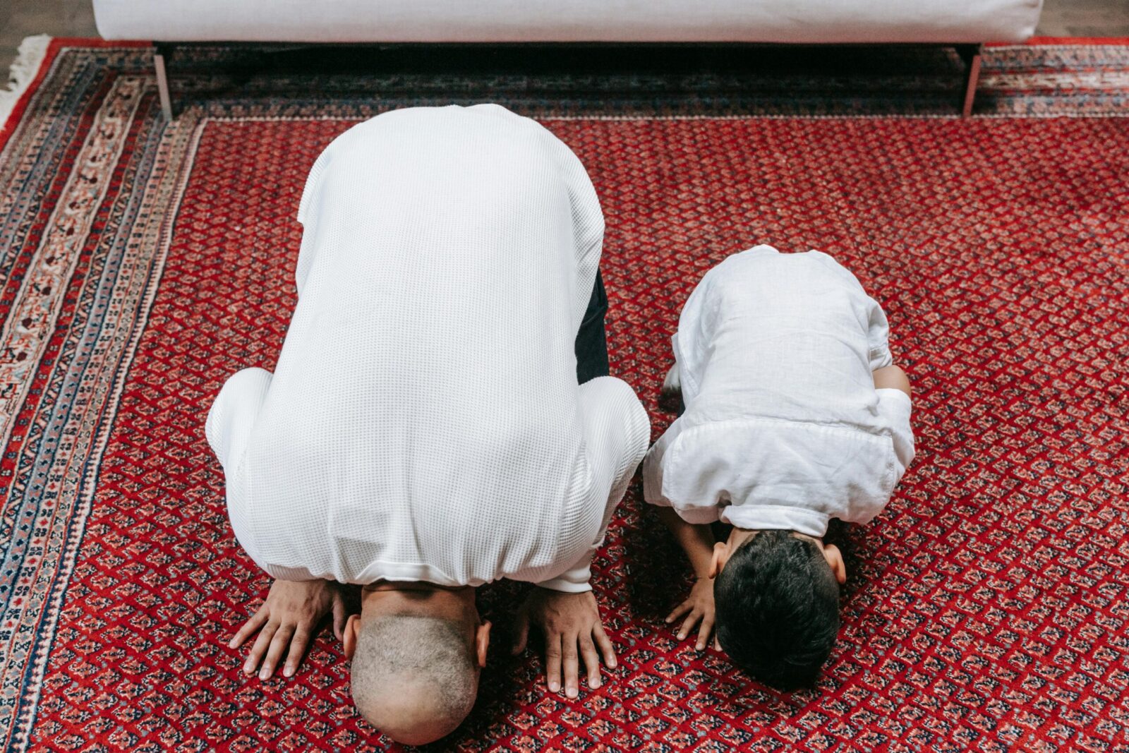 A father and son bowing in prayer on a traditional rug indoors, displaying religious devotion.
